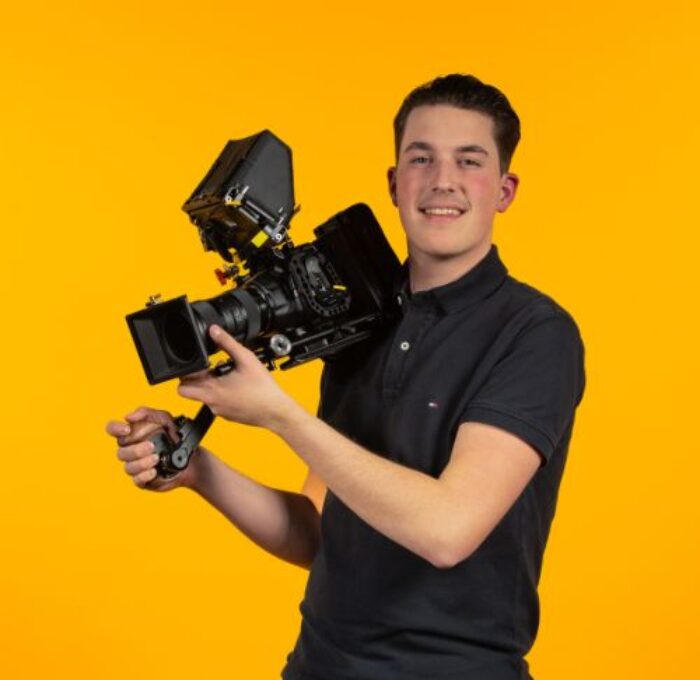 Hugo Brandwagt on a yellow background with a filmcamera on his right shoulder.