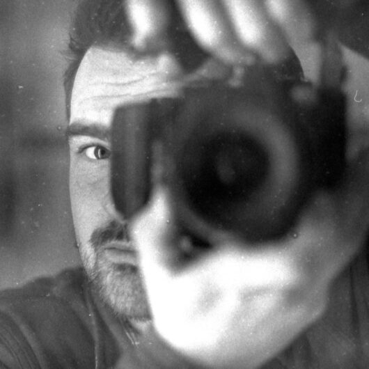 Old action photo of Nanko Goeting behind the lens of a DSLR camera with lens. He holds the camera up to his face. It is a black-and-white photo.