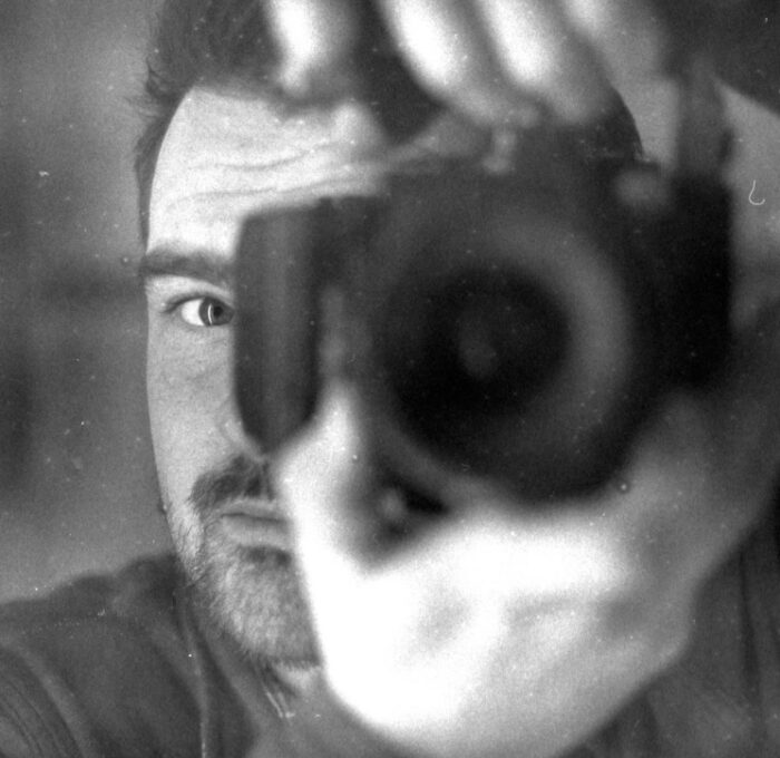 Old action photo of Nanko Goeting behind the lens of a DSLR camera with lens. He holds the camera up to his face. It is a black-and-white photo.